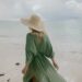 Back view of unrecognizable female tourist wearing straw hat and summer dress standing on seashore in cloudy day